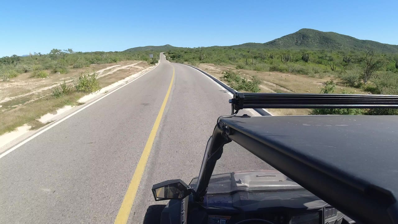 Empty paved road viewed from a moving black vehicle in dry green scrubland.