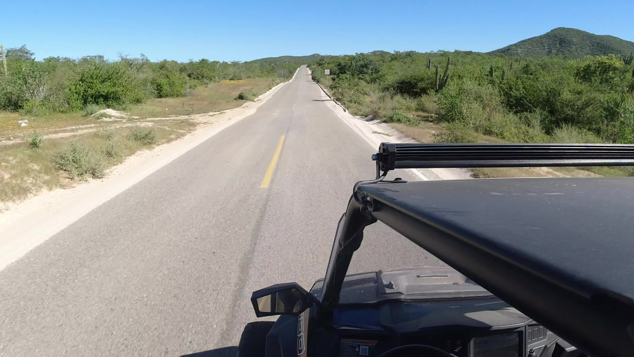 The Los Cabos highway continues straight ahead with the black SUV foreground shifted along the lane, dense green brush to the right, sandy roadside patches to the left, distant hills, and no visible people.
