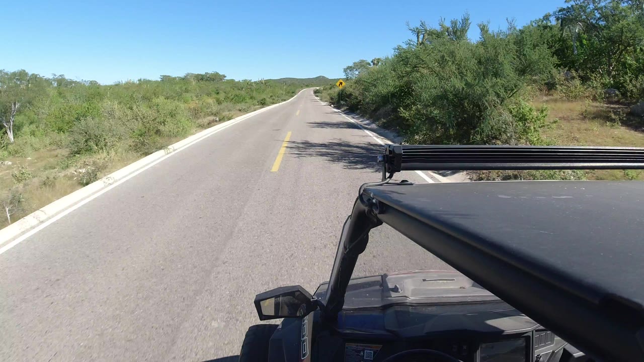 Vehicle-mounted view down a straight Los Cabos road with green brush on both sides, blue sky overhead, black car hardware in the foreground, and no visible people.