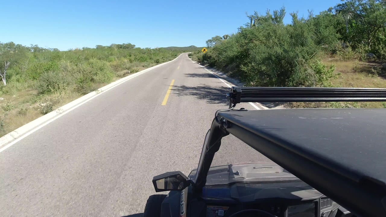 A high vehicle perspective looks along the Los Cabos road as scrub vegetation lines both shoulders, with the black roof and mount occupying the lower right and no visible people.