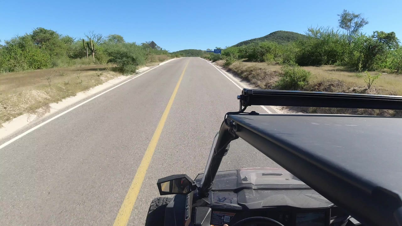 Opening Los Cabos road view with a centered yellow line, dry roadside brush, green trees, distant rounded hill, vehicle roof visible at lower right, and no visible people.