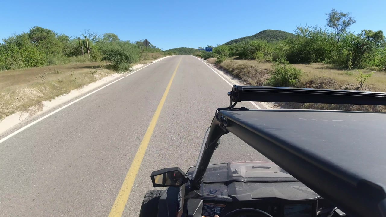 Los Cabos road scene one second in, with the vehicle roof and support bar in the foreground, low green vegetation on both sides, a rounded hill ahead, and no visible people.