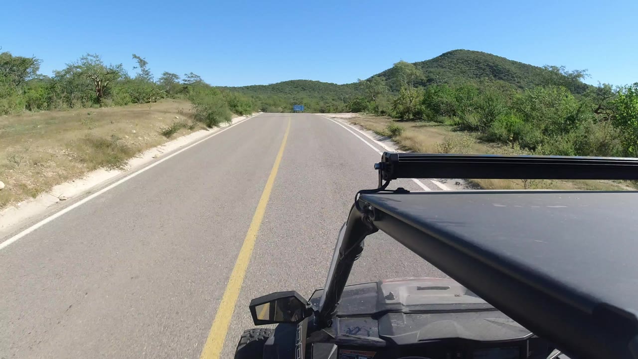 Forward vehicle view on a two-lane road in Los Cabos, Mexico, with a yellow center line, green roadside trees, distant low hills, clear blue sky, and no visible people.