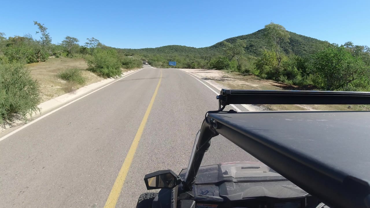 Road curving slightly right with desert shrubs, hills, and part of a vehicle in view.