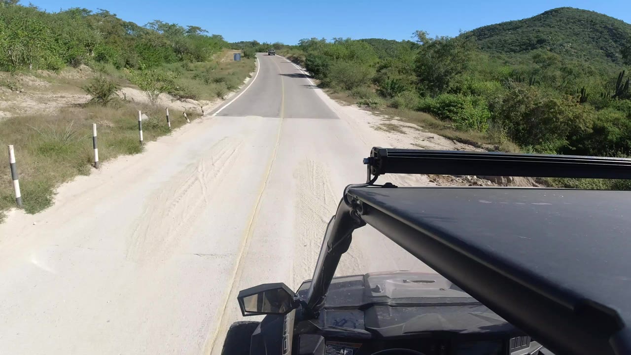 Forward view from a vehicle on a paved road in Los Cabos, Mexico, with pale roadside sand, green shrubs, low hills ahead, and no visible people.