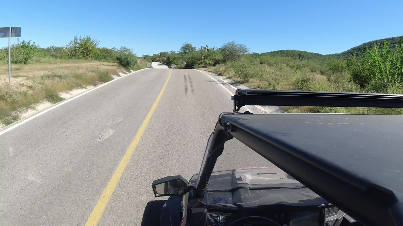 Forward road view in Los Cabos, Mexico, with the vehicle roof and side mirror in the foreground, low green brush beside the pavement, distant hills, and no visible people.