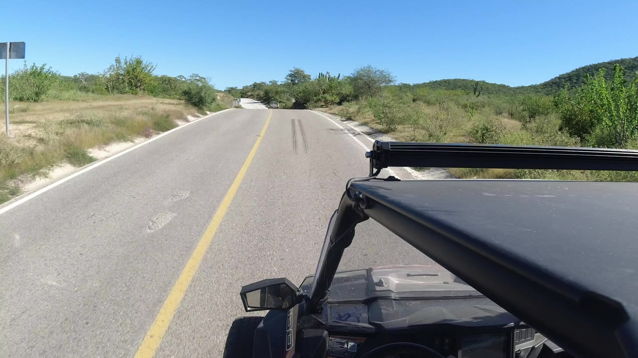 The road remains centered in Los Cabos, Mexico, with a black vehicle roof edge at lower right, dry grasses and green shrubs on both shoulders, distant hills, and no visible people.
