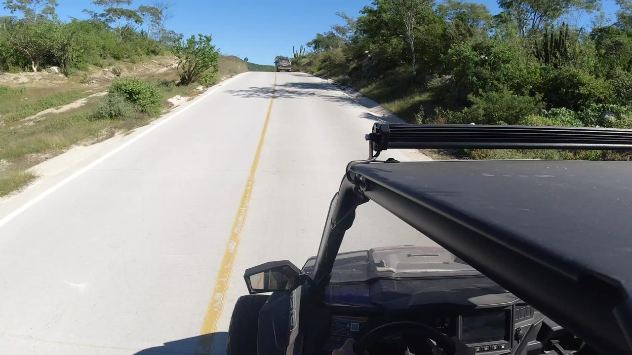 Forward vehicle-mounted view on a pale road in Los Cabos, Mexico, with scrub trees on both sides, a distant vehicle ahead, blue sky, and no visible people.