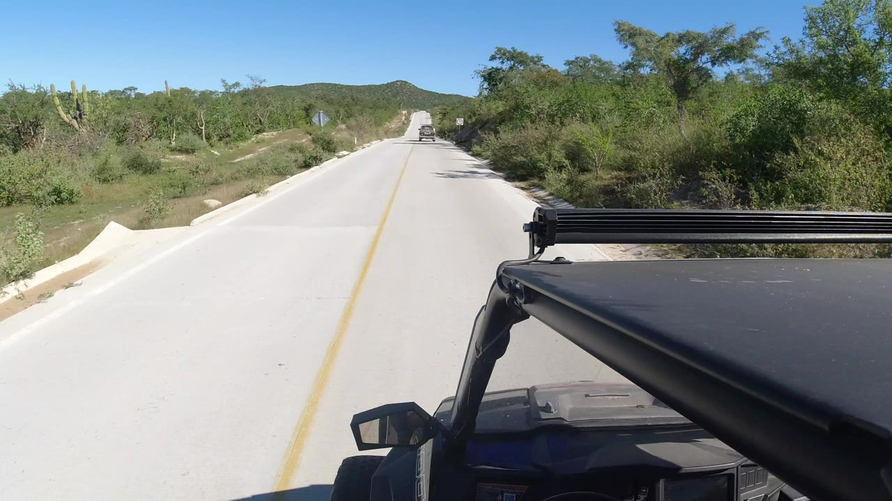 Opening frame of a Los Cabos road with green brush on both sides, low hills ahead, part of a black vehicle in the foreground, and no visible people.
