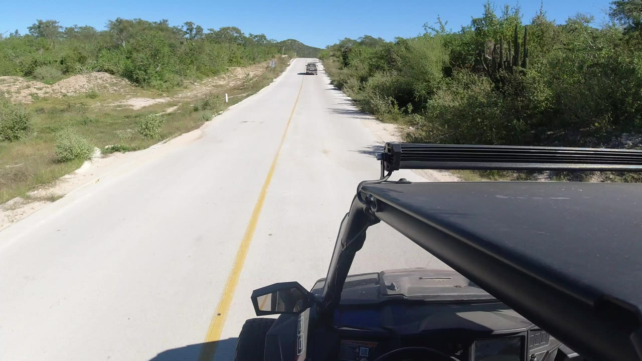 Start of a Los Cabos road view from an open-top vehicle, with a yellow center line, sandy shoulders, green desert brush, distant low hills, and no visible people.