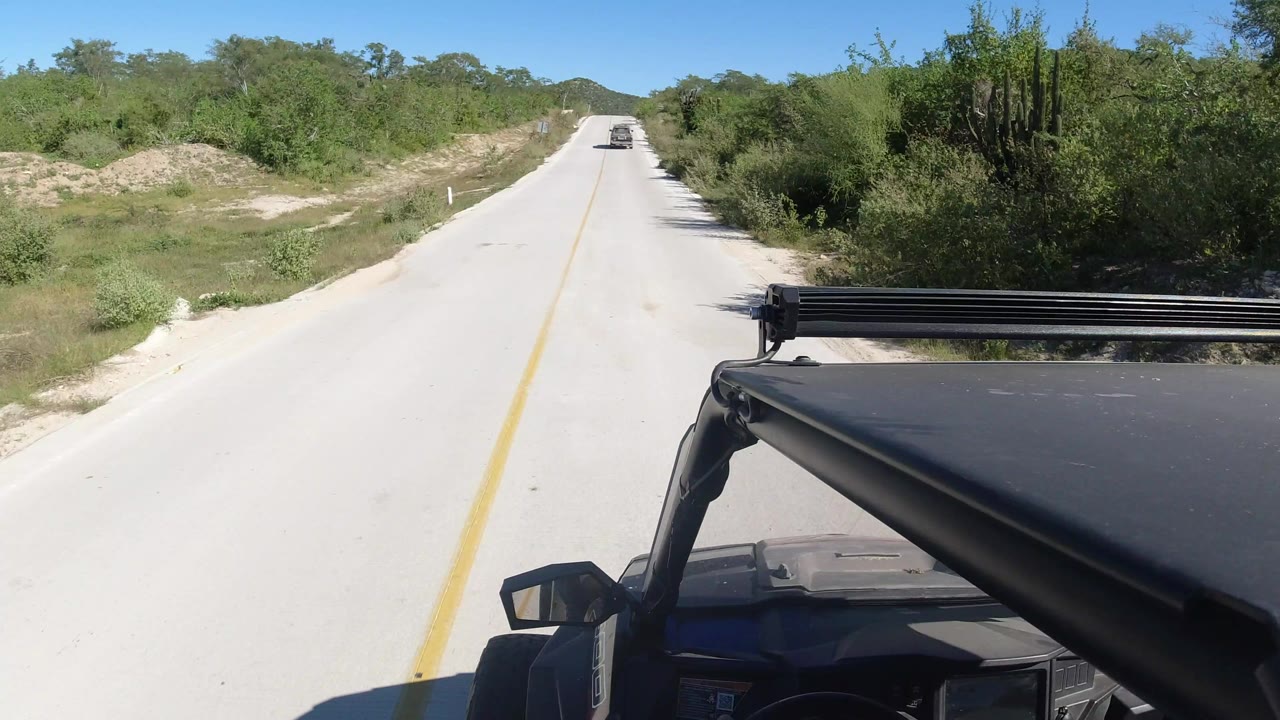 A Los Cabos paved road continues straight ahead, with the open-top vehicle foreground on the right, sandy roadside vegetation, clear blue sky, and no visible people.