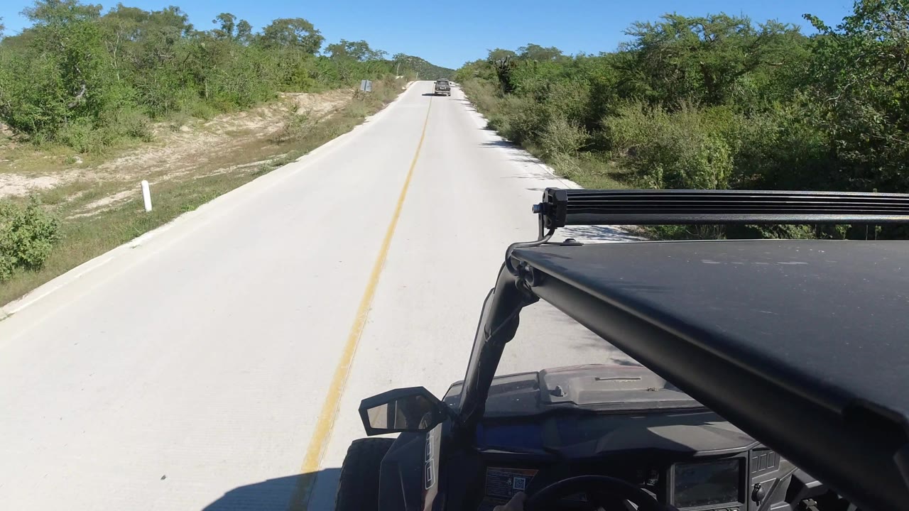 Bright road ahead with scrubland and the vehicle roof visible at right.