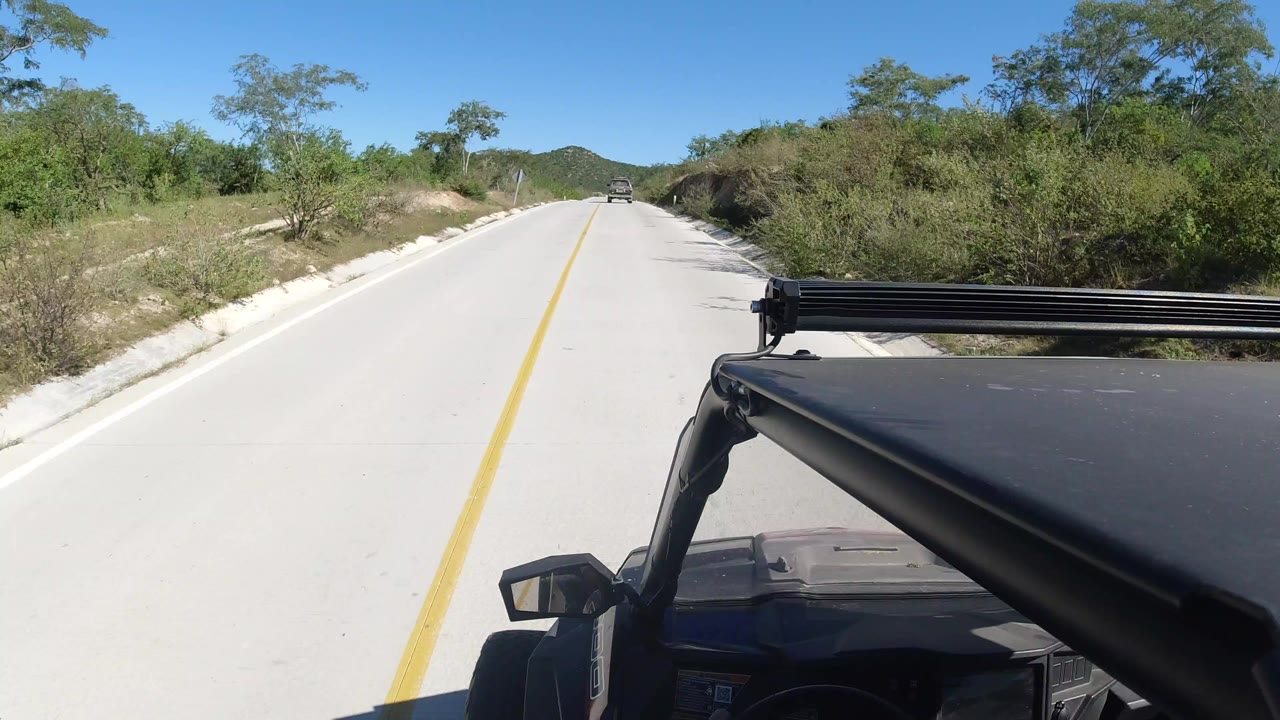 Opening frame of a Los Cabos road view with a pale paved lane, yellow center line, distant vehicle, green roadside brush, low hills, blue sky, and no visible people.