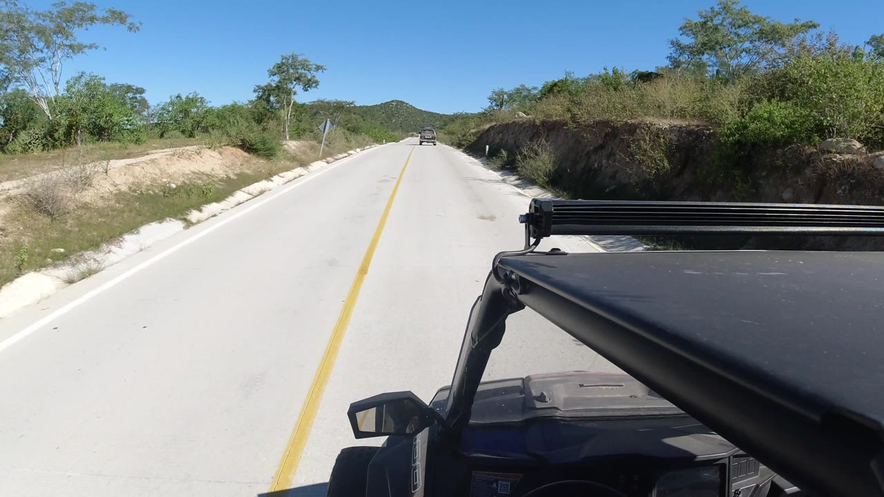 Road POV passing a sandy shoulder and roadside brush under blue sky.