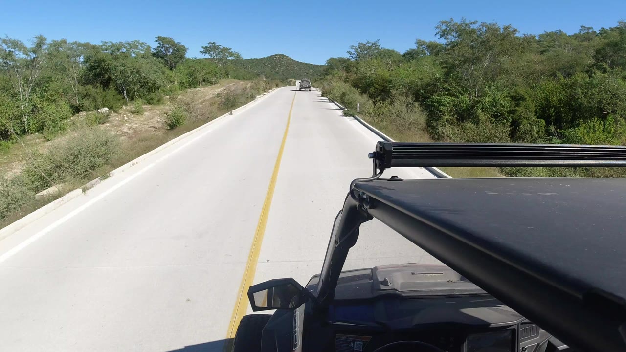 Opening frame of a Los Cabos road POV, with a yellow centerline, green roadside brush, distant low hills, part of the vehicle in the lower right, and no visible people.