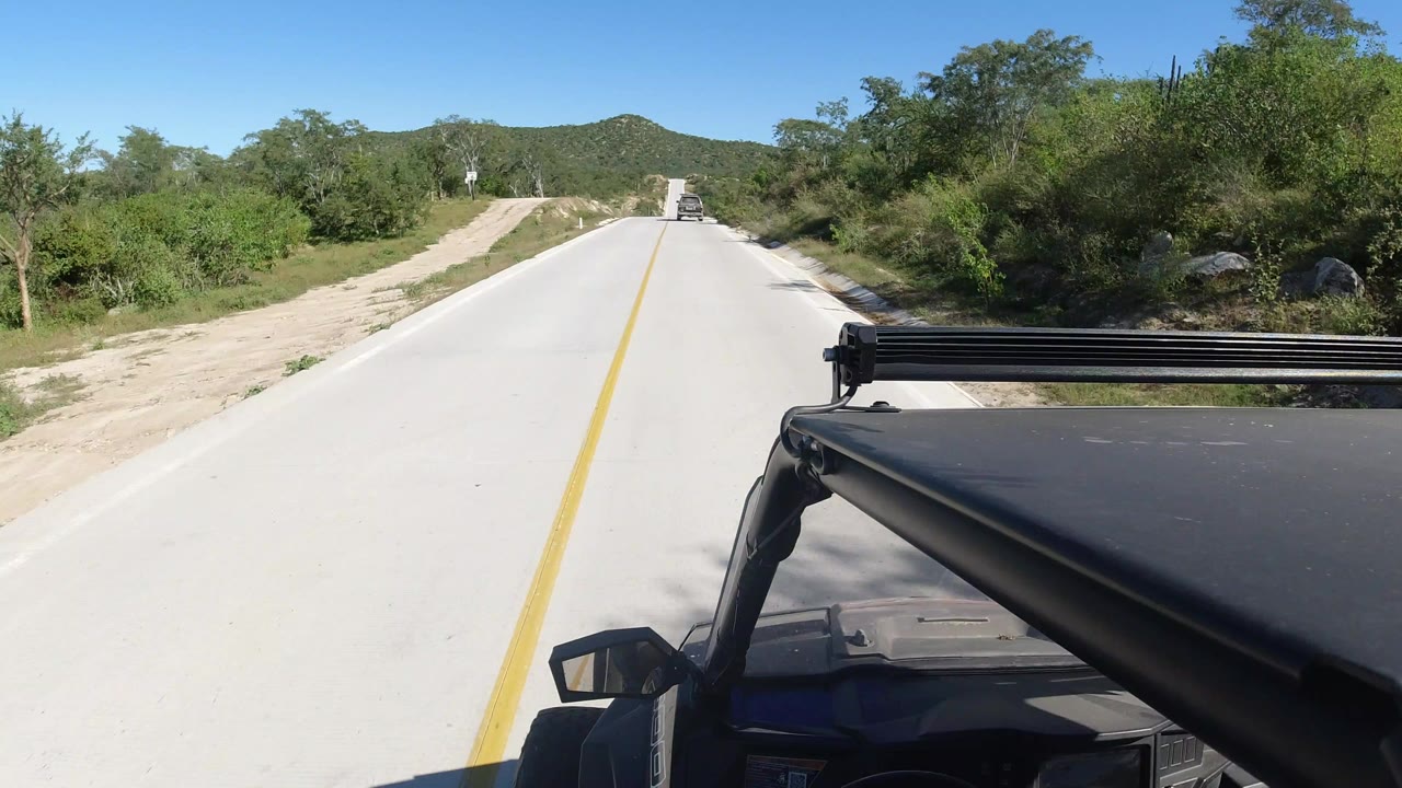 Forward view down a sunny paved road from an open black vehicle.