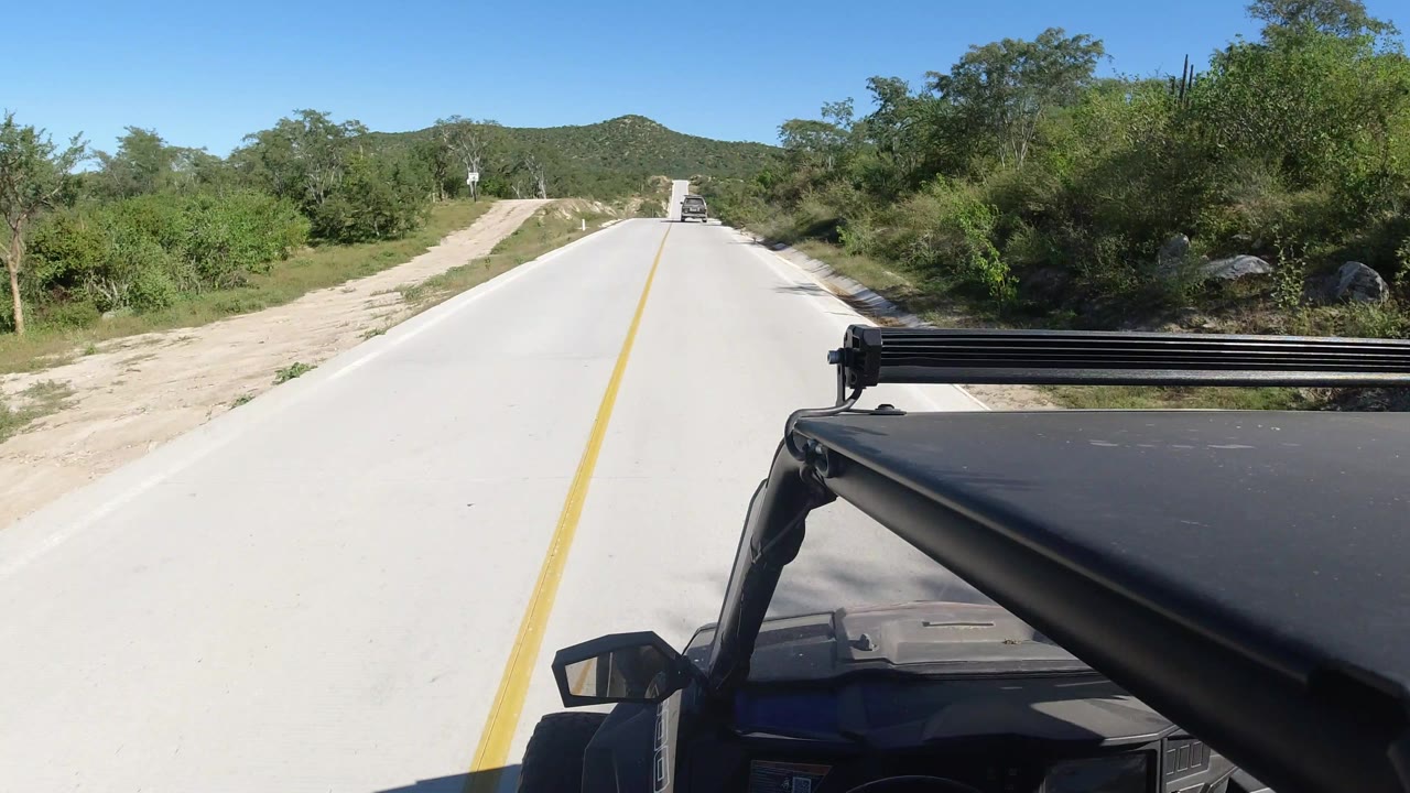 Los Cabos road POV with the yellow centerline leading toward low green hills, dense brush at right, sandy shoulder at left, and no visible people.