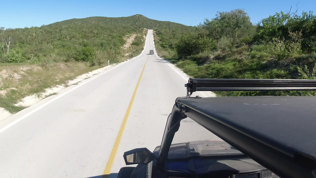 Forward-facing view of a pale two-lane highway in Los Cabos, Mexico, with green shrubland on both sides, low hills ahead, part of the vehicle in the lower right, and no visible people.