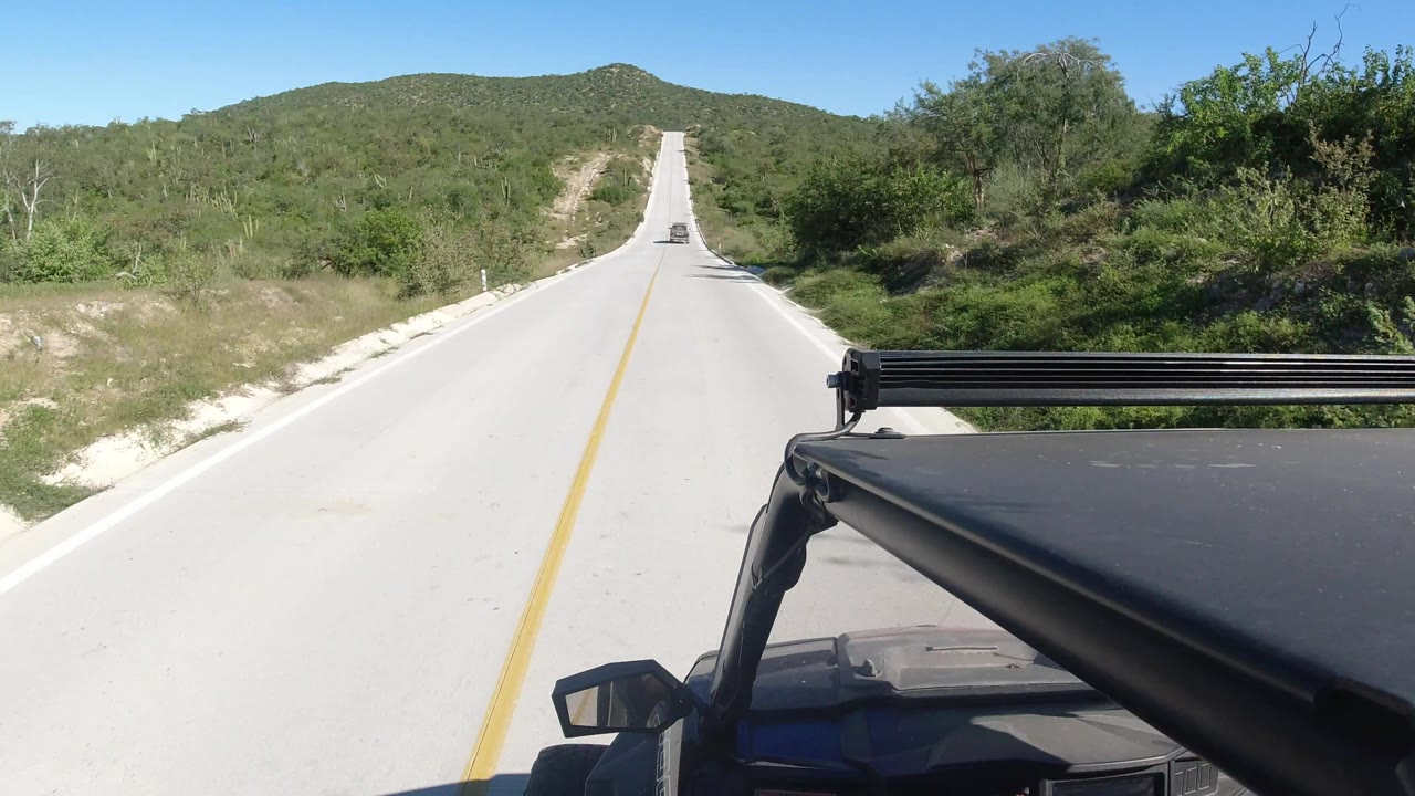 A centered Los Cabos highway view shows the yellow line stretching uphill, thick green bushes along the shoulders, the filming vehicle's roof rack and mirror at right, and no visible people.