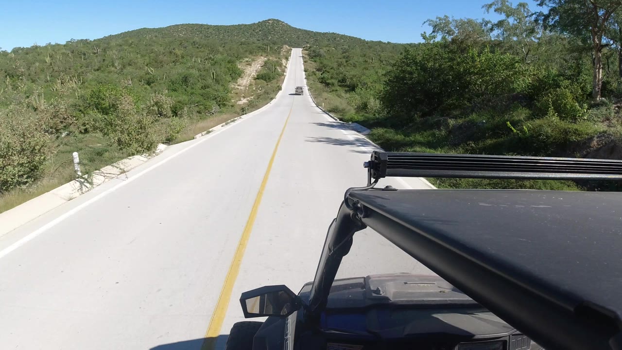 The Los Cabos road rises toward green hills under a clear sky, with a small vehicle far ahead, roadside brush close to both lanes, vehicle bodywork at lower right, and no visible people.
