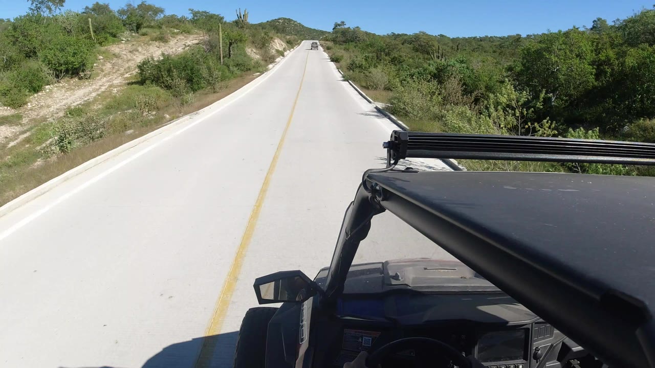 Forward view from an open Jeep on an empty concrete road through green brush.