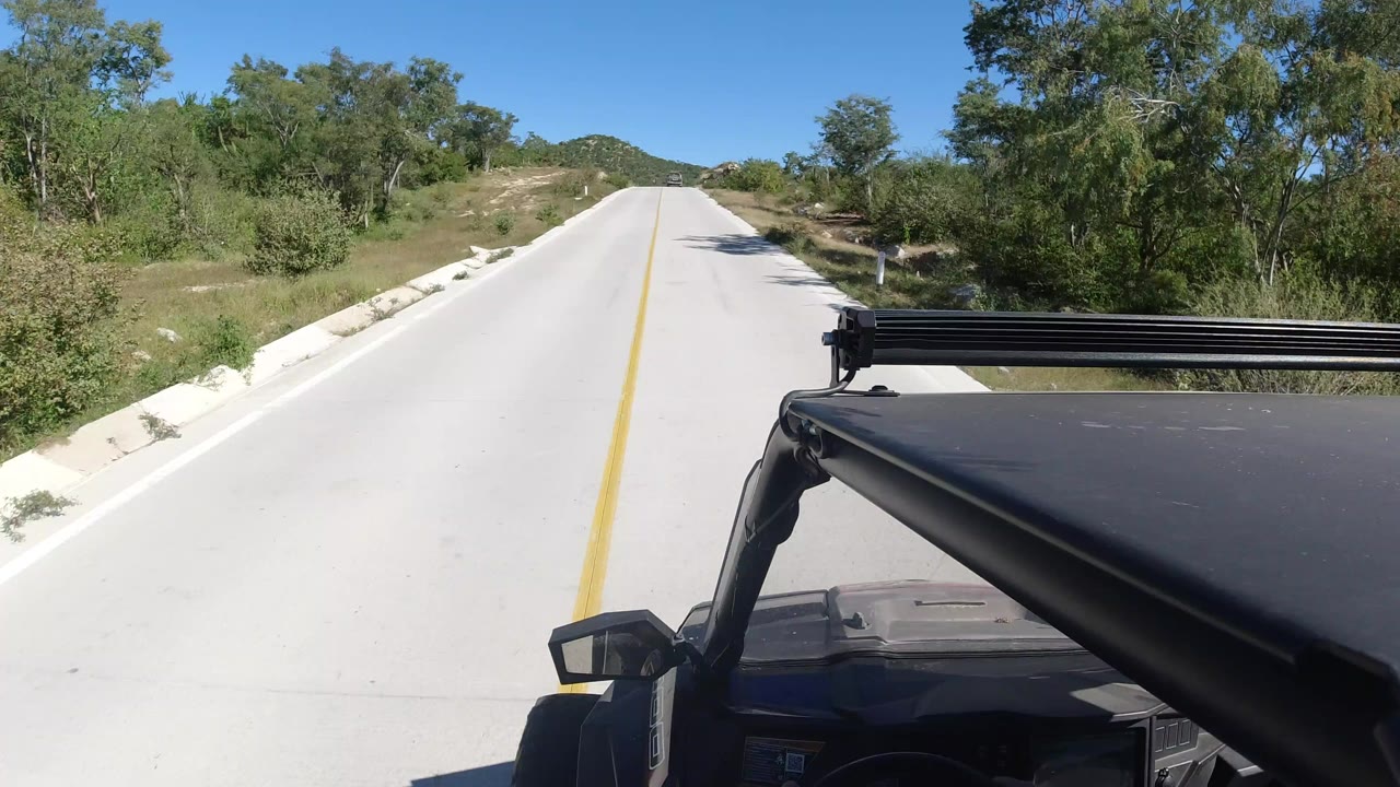 Forward vehicle view on a pale rural road in Los Cabos, Mexico, with scrub trees on both sides, low hills ahead, blue sky, and no visible people.