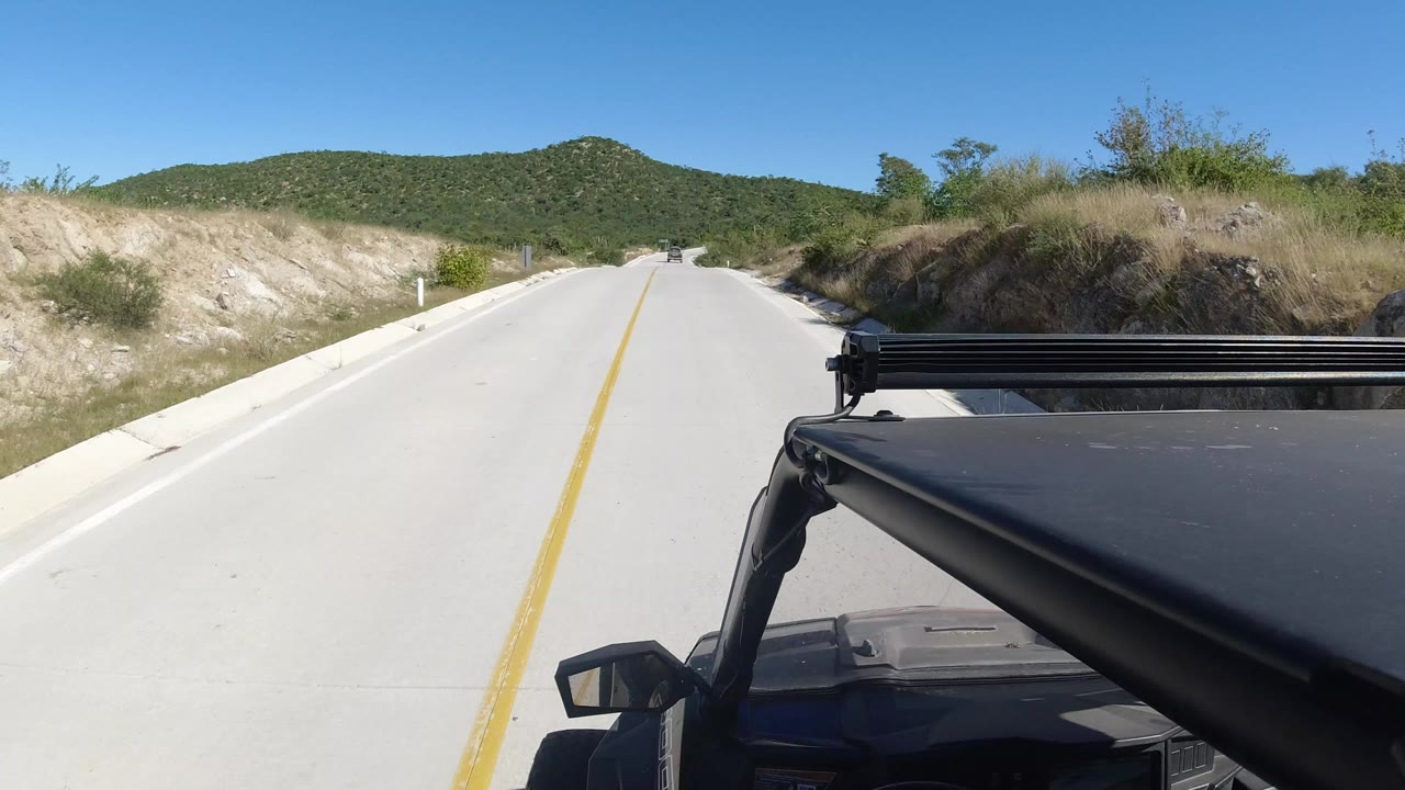 Opening frame of a Los Cabos, Mexico road seen from a dark vehicle, with a yellow center line, dry roadside banks, green hills ahead, bright sun, and no visible people.