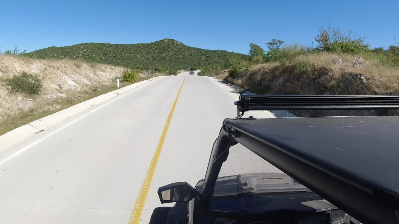 At one second, the Los Cabos road continues straight ahead with the dark vehicle roof and rack in the foreground, scrub-covered banks beside the pavement, distant hills, and no visible people.
