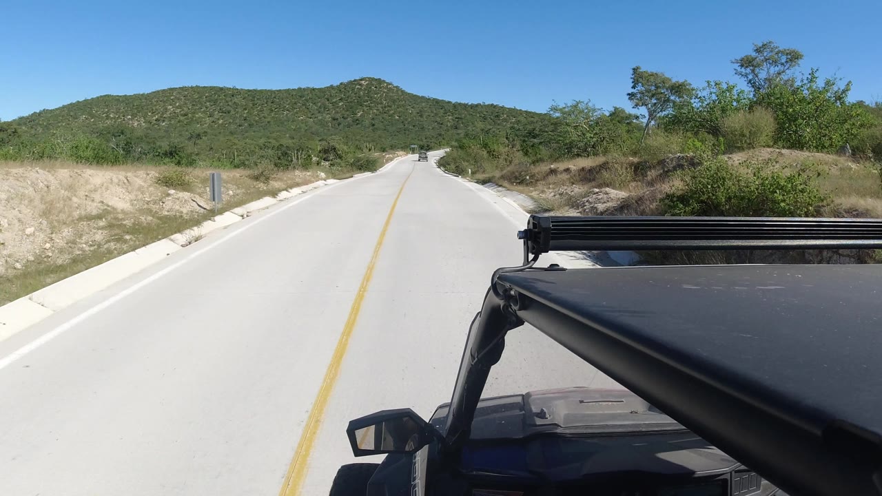 Curving road ahead with a dark vehicle roof in the foreground and hills beyond.