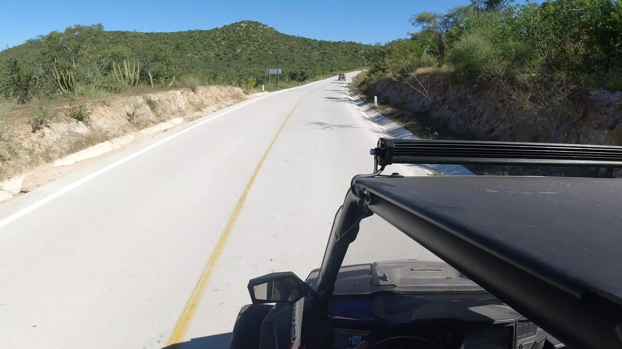 Forward road view from a black vehicle on a sunny hillside road.