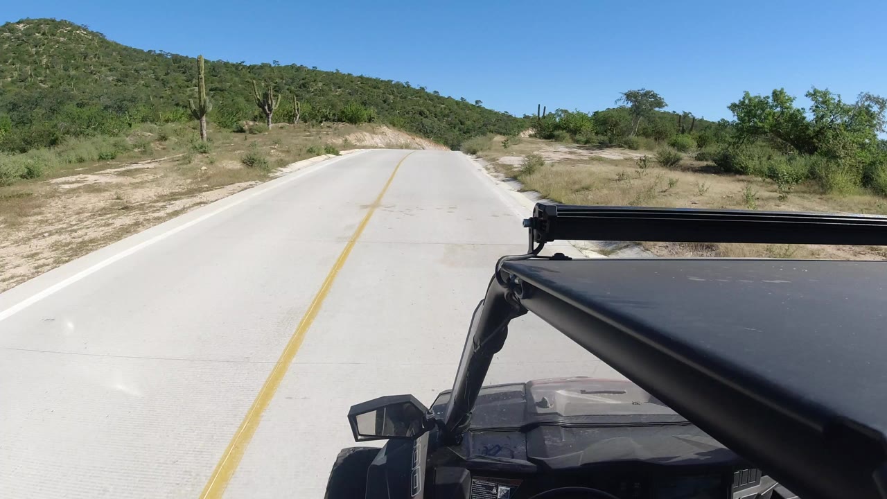 Opening vehicle POV on a pale two-lane road in Los Cabos, Mexico, with a yellow center line, green desert hills, tall cacti, and no visible people.