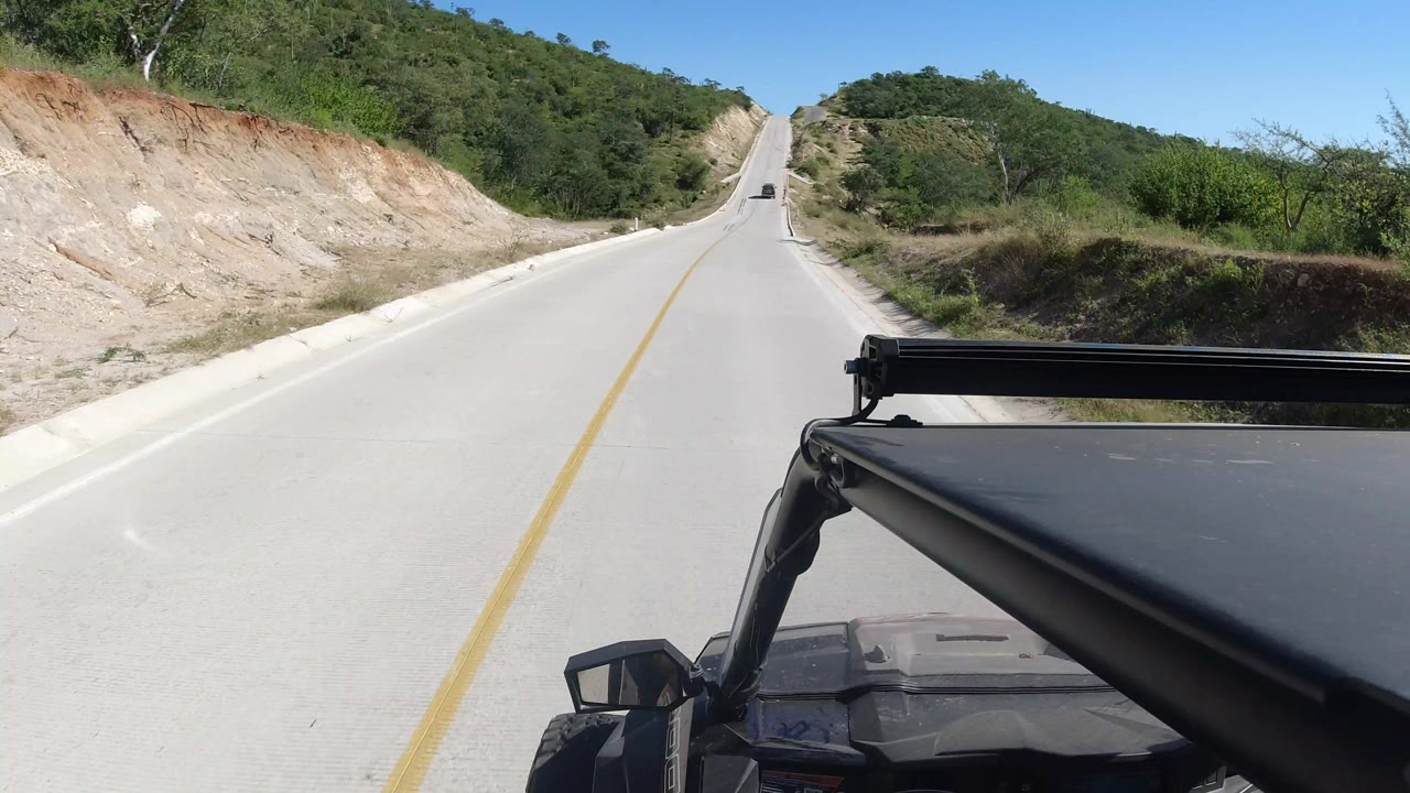 Forward view from a vehicle on a pale road between dry hills.