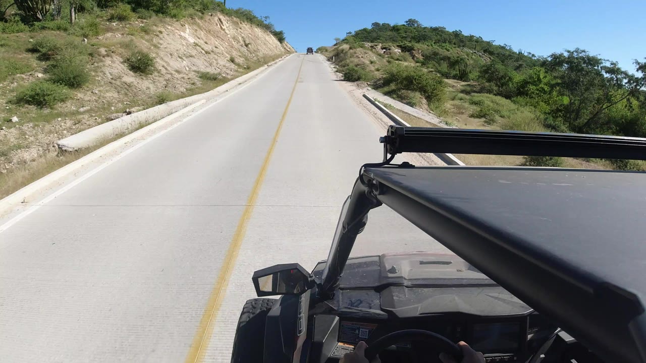 Forward view from a vehicle on a Los Cabos highway, with a pale road climbing between cut hills, green shrubs, blue sky, and no visible faces.