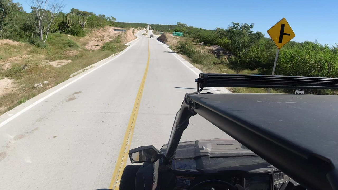 The Los Cabos road stretches uphill ahead as the Land Rover foreground remains at the lower right, with dry earth shoulders, green vegetation, a yellow directional sign, and no visible people.