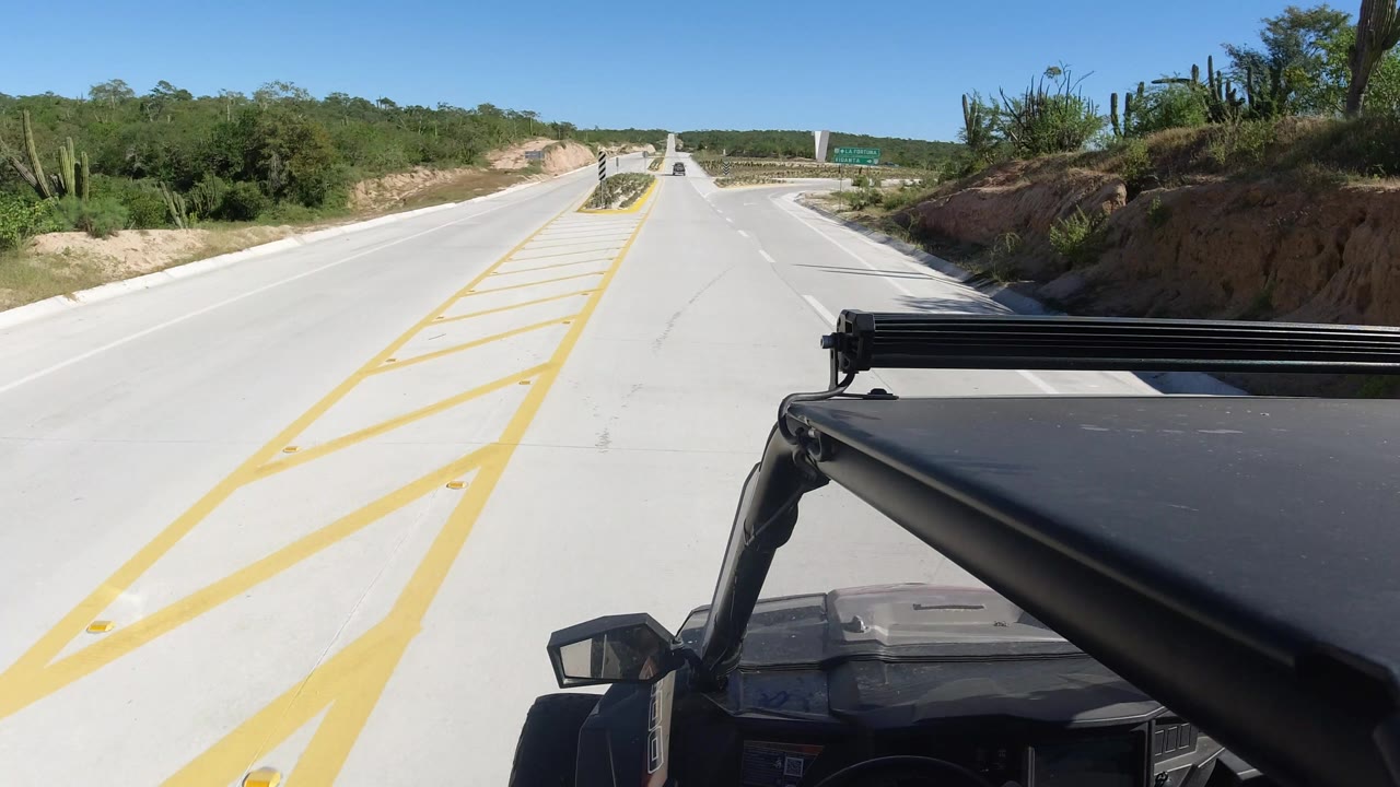 Los Cabos roadway seen past the black vehicle edge, with a sandy cut bank on the right, low green vegetation, distant signs, and no visible people.