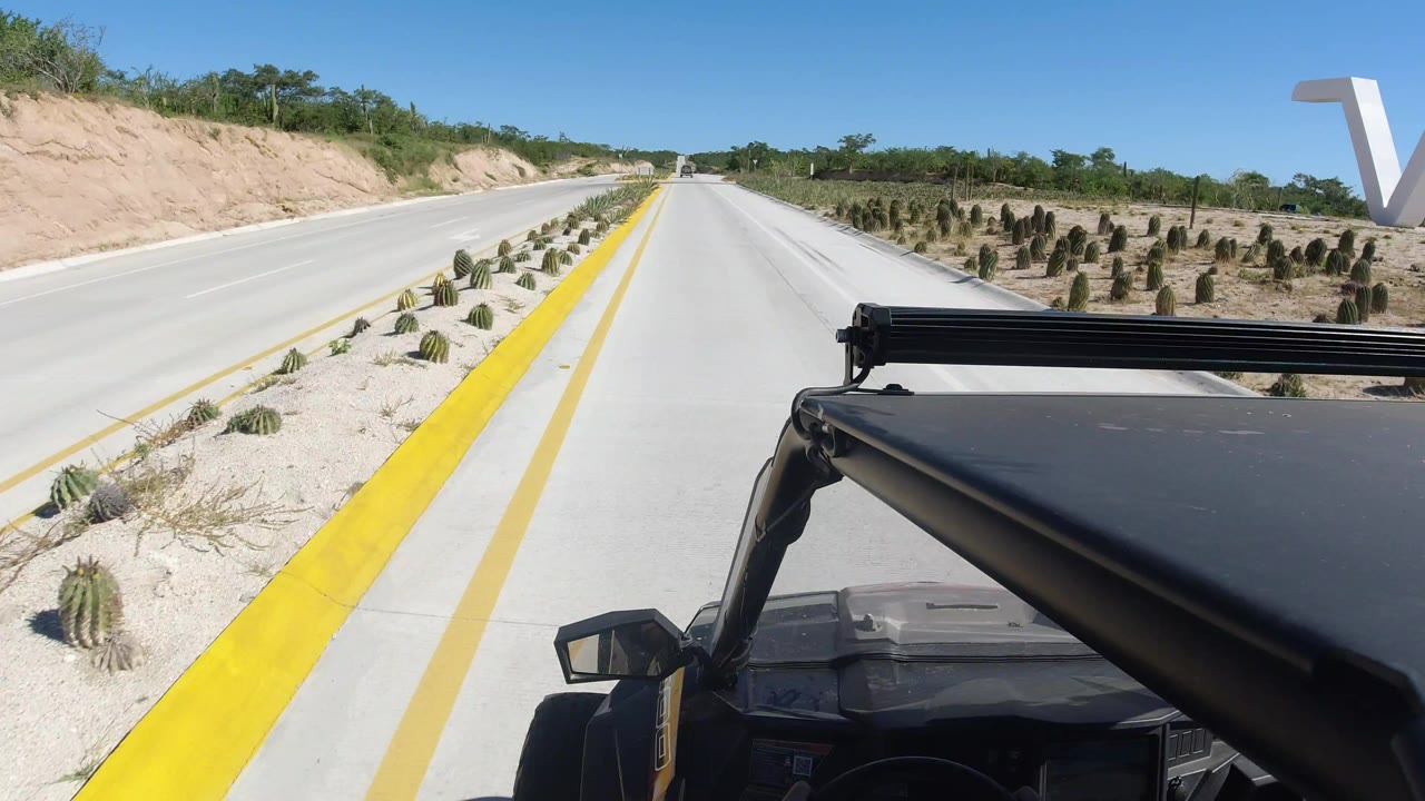 Roof-mounted view from a jeep on a quiet Los Cabos road, with yellow curb lines, cactus plantings, scrubby banks, blue sky, and no visible people.