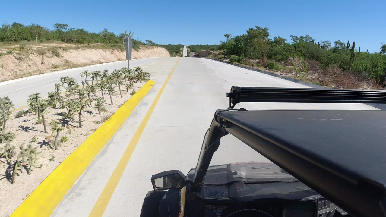 Forward-facing SUV view on a pale highway in Los Cabos, Mexico, with a yellow curb, sparse roadside plants, dry banks, blue sky, and no visible people.