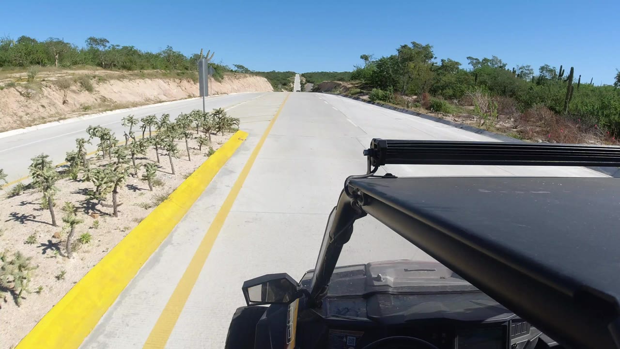 Moving SUV perspective on a Los Cabos highway with the yellow curb closer at left, dry roadside planting beds, green brush, and no visible people.