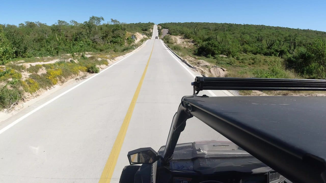 A forward view from a black vehicle on an empty paved road in Los Cabos, Mexico, with a yellow center line, low green brush, sandy shoulders, and no visible people.