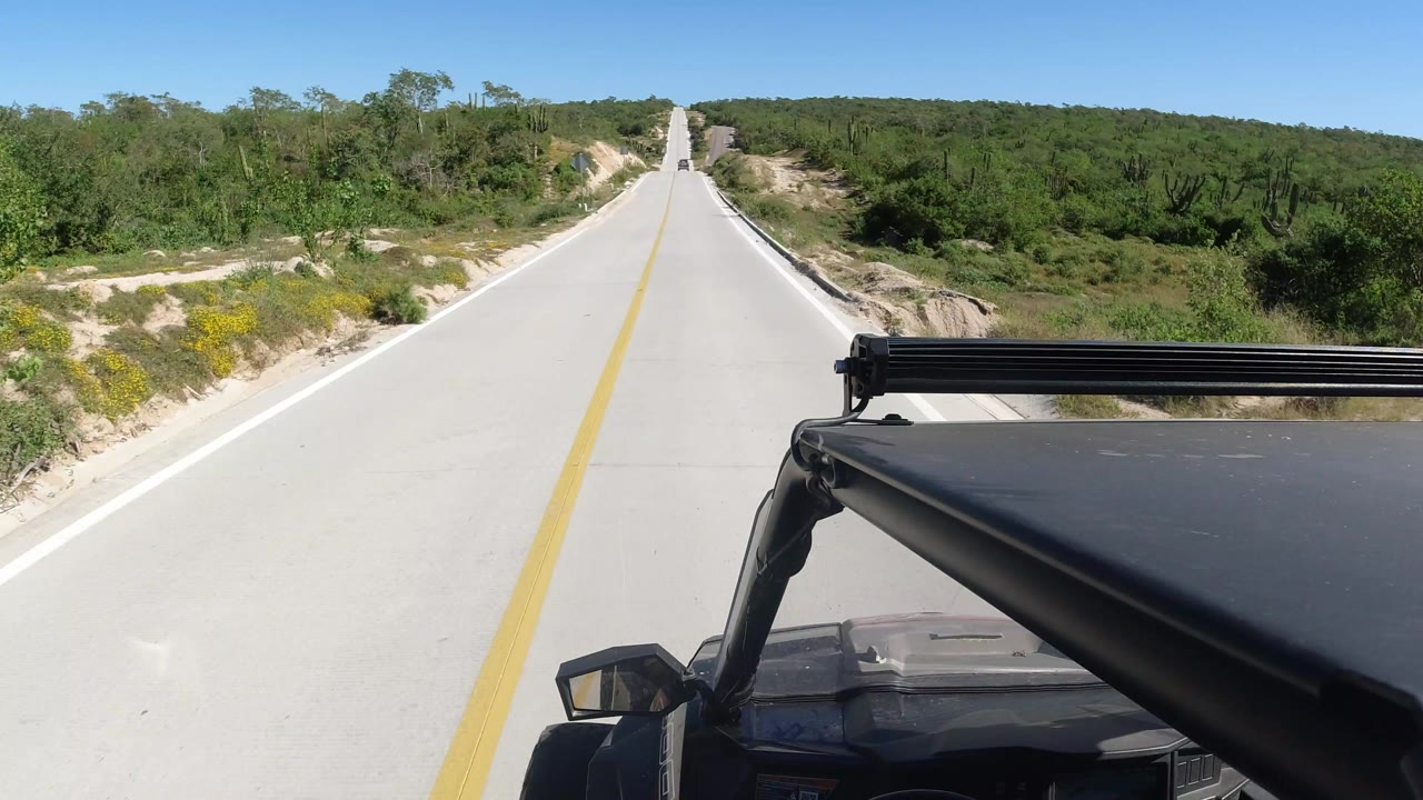 The road stays centered ahead in Los Cabos, Mexico, while the black vehicle hood and roof rack fill the lower right foreground and scrubby vegetation lines both sides with no visible people.