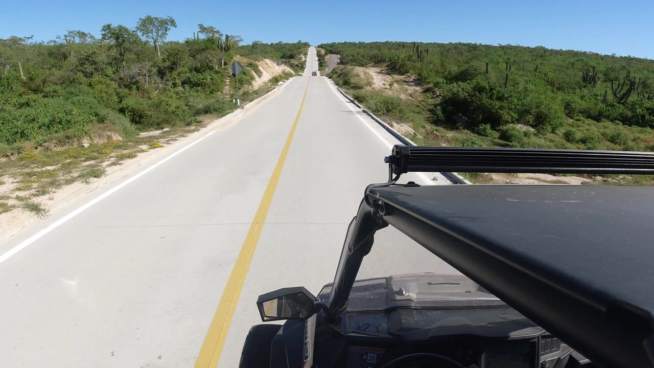 Paved road curving past brush with a black vehicle corner visible.