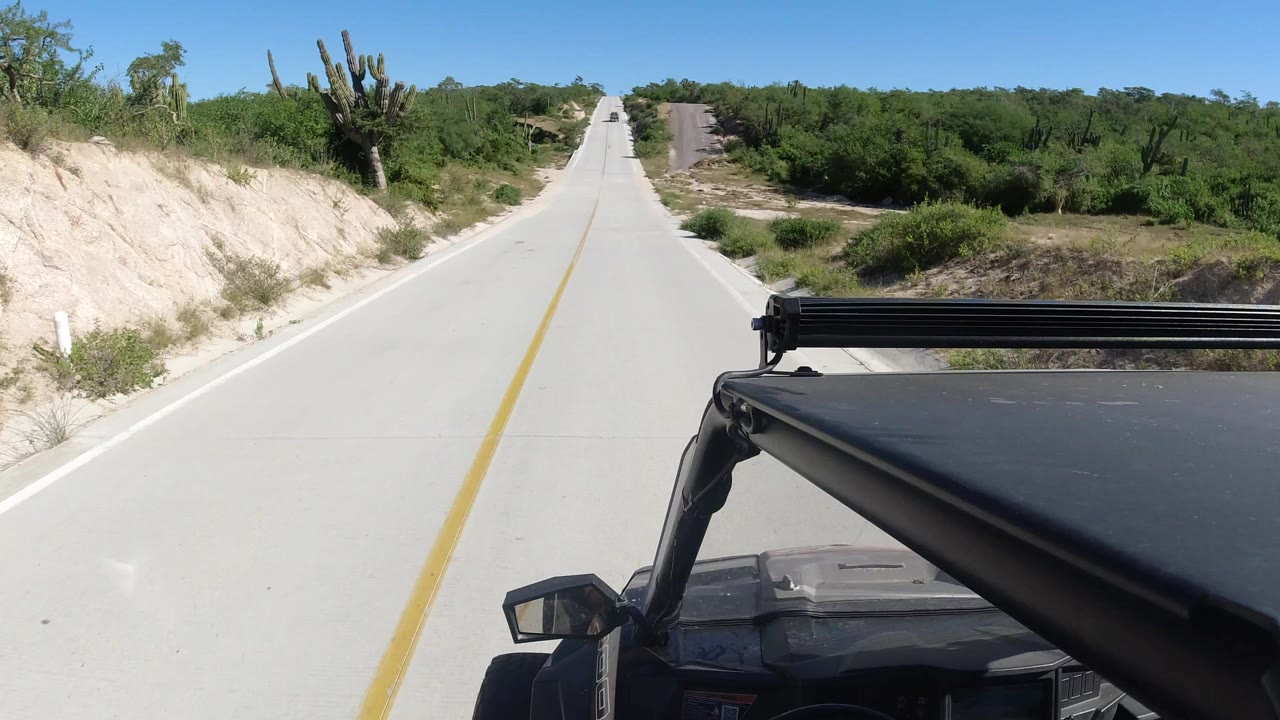 Opening frame from Los Cabos, Mexico shows a pale two-lane road ahead with a yellow centerline, sandy banks, green brush, and part of a black SUV visible in the foreground, with no visible people.