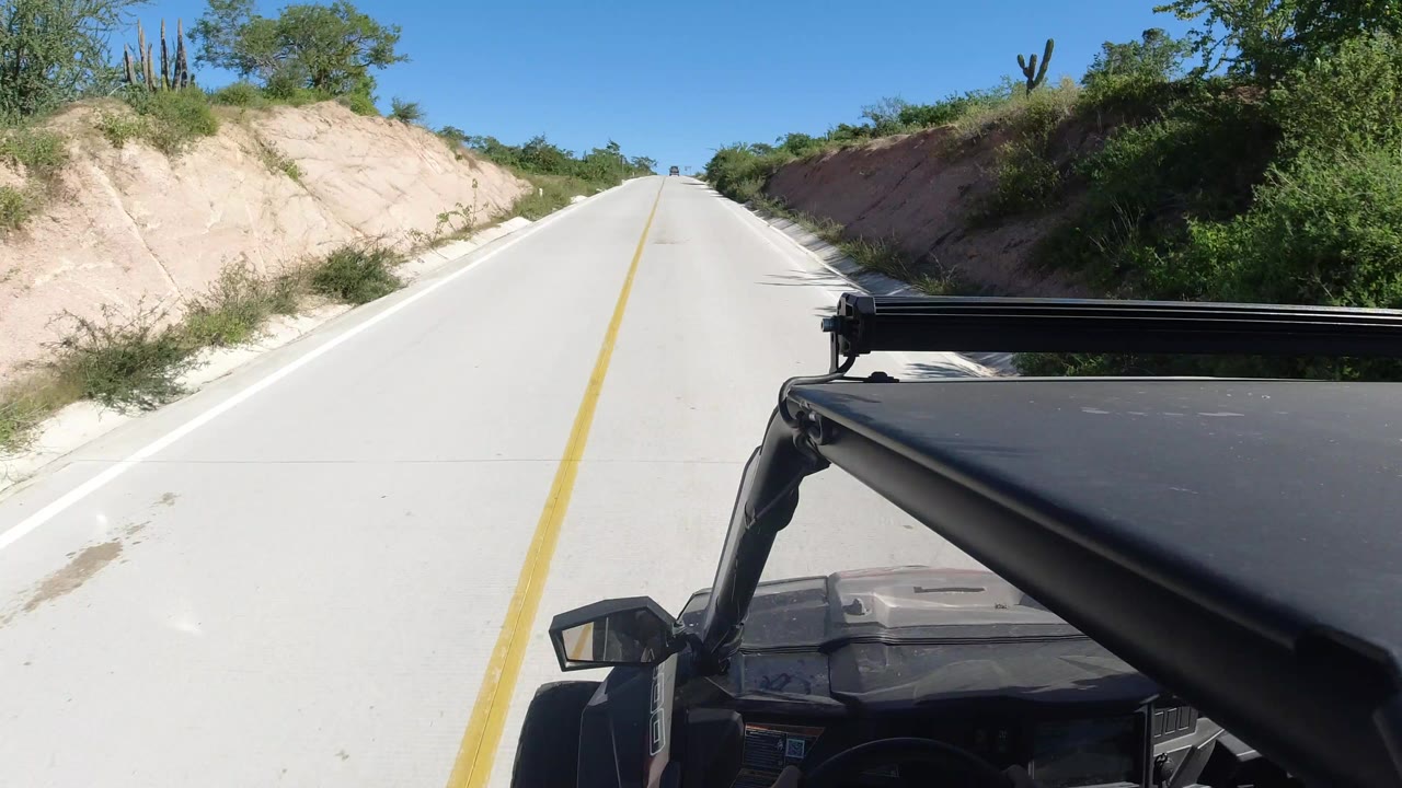 Opening frame of a Los Cabos road seen from a black vehicle, with a yellow center line, sandy cut banks, green shrubs, cactus silhouettes, and no visible people.