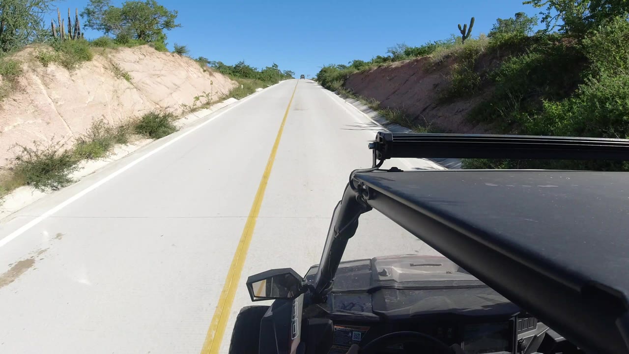 Los Cabos driving view one second in, with the pale road rising ahead, the black vehicle structure at lower right, green roadside scrub, sandy shoulders, and no visible people.