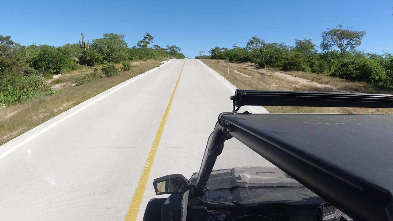 Forward vehicle POV on a straight pale road in Los Cabos, Mexico, with black vehicle hardware in the foreground, scrub vegetation along both shoulders, and no visible people.