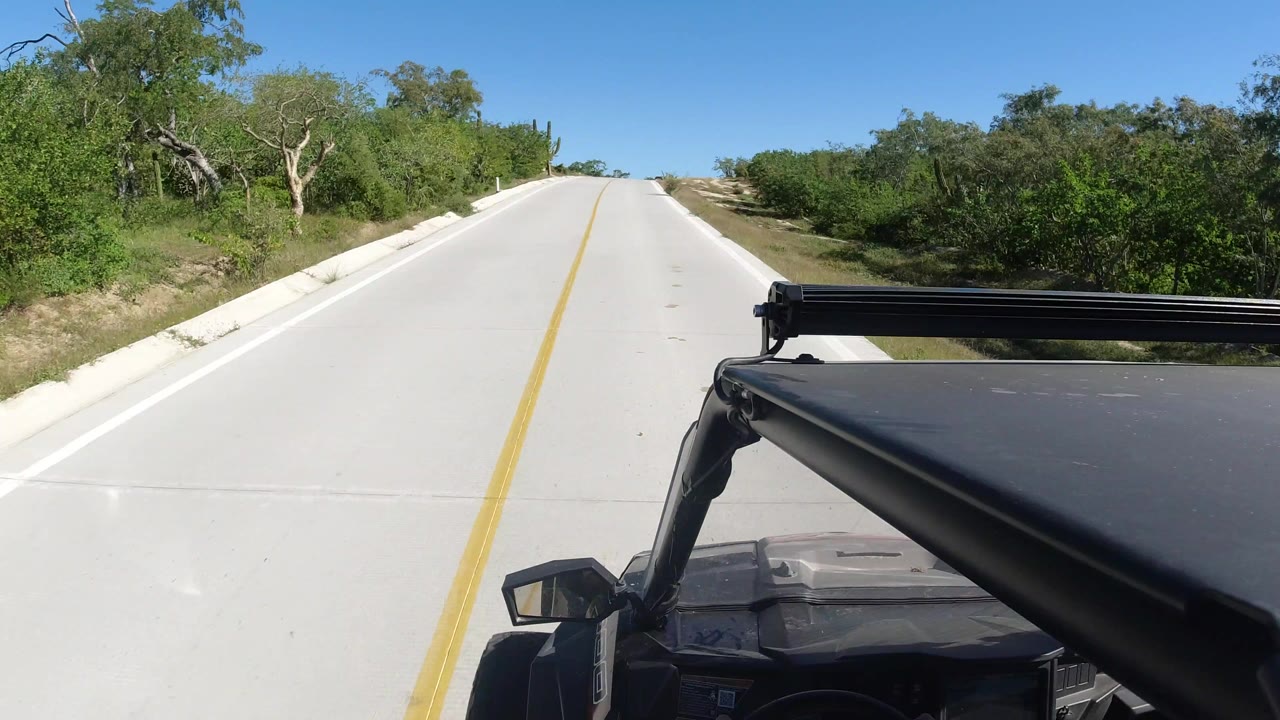 Start of the Los Cabos road clip showing a pale concrete lane, yellow centerline, green scrub on both sides, blue sky, and no visible people.