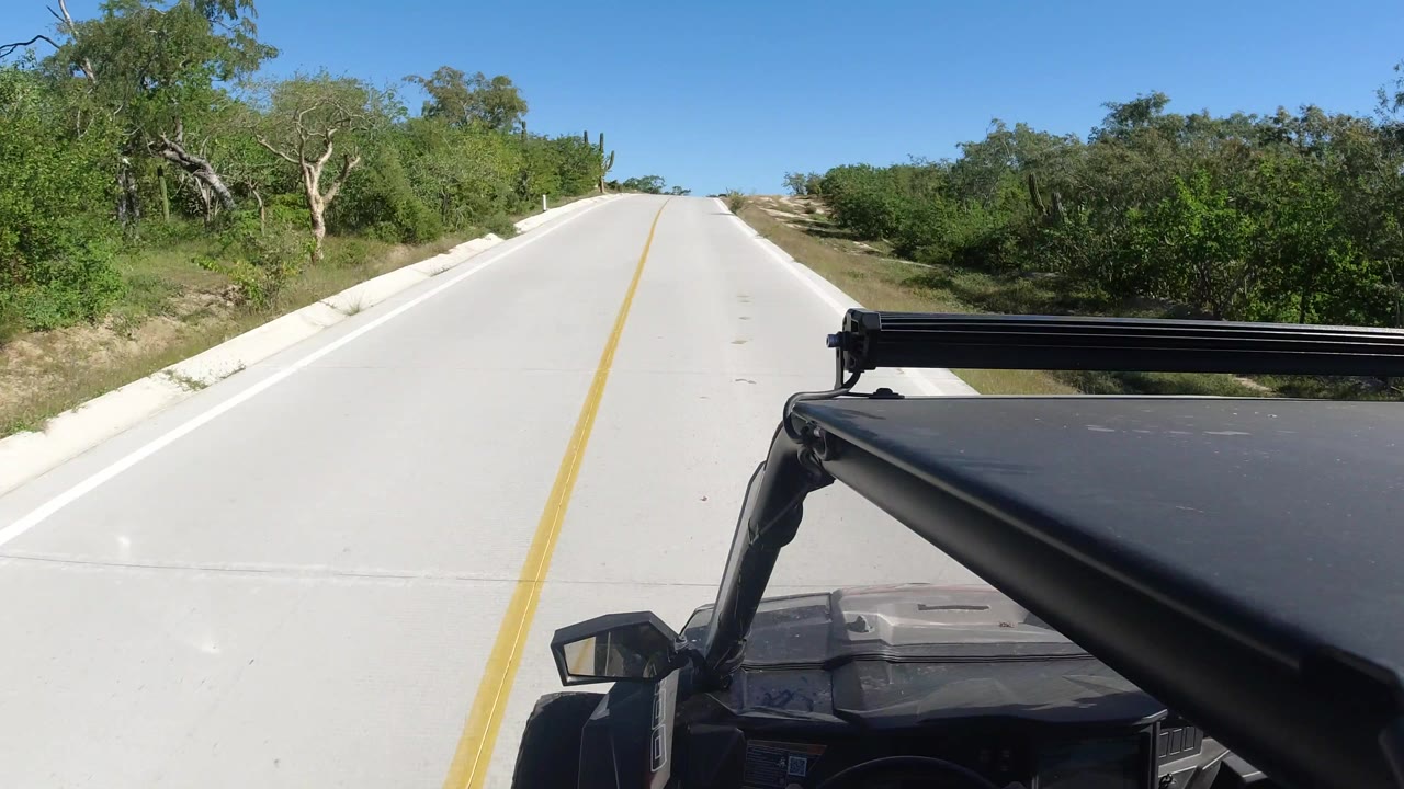 Los Cabos road view one second in, with the centered yellow line leading past dense green brush and no visible people.