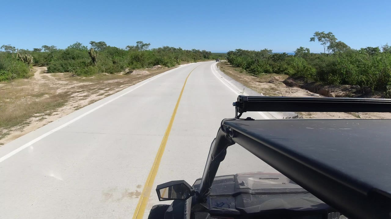 Start of the Los Cabos, Mexico road clip with a pale concrete lane, yellow center stripe, green brush on both sides, black vehicle hardware in the foreground, and no visible people.