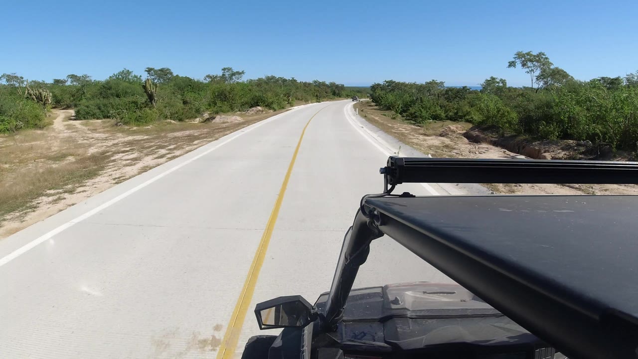 Los Cabos, Mexico roadway seen from a vehicle-mounted angle, with the yellow stripe leading ahead, low shrubs beside the road, a clear blue sky, and no visible people.
