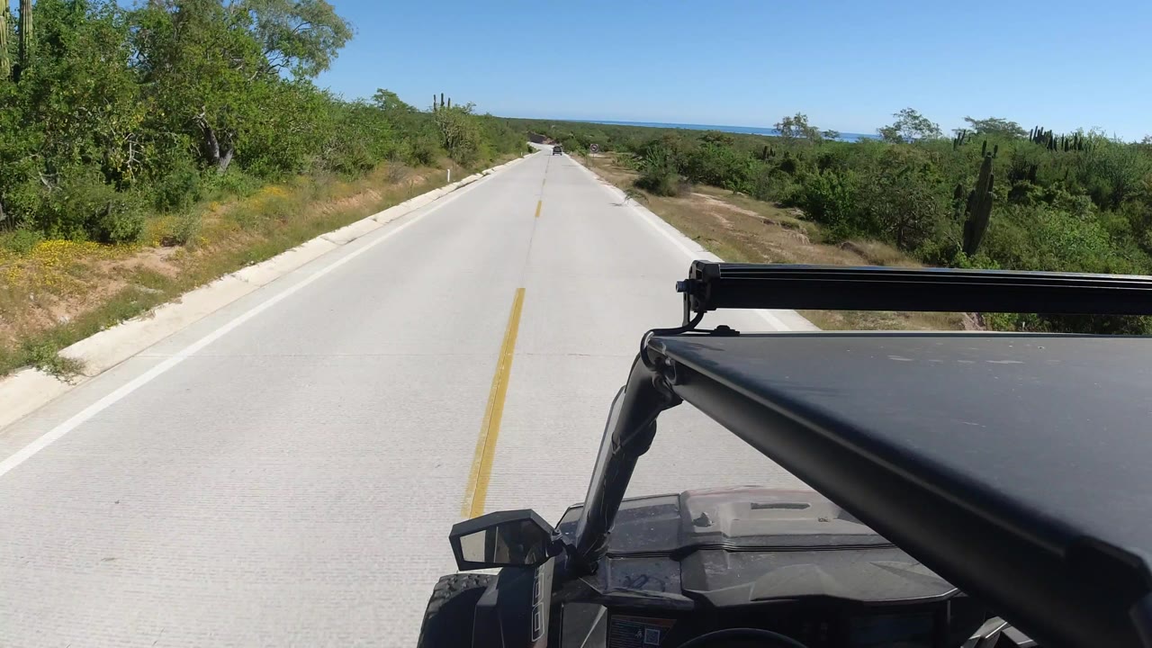 The road remains centered as the vehicle continues through Los Cabos scrubland, with dry shoulders, green trees, cactus, a low horizon, and no visible people.